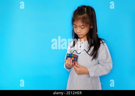 Une jeune fille asiatique mignonne joue avec un cube de Rubik, s'amusant tout en apprenant. Fond bleu clair Uni. Banque D'Images