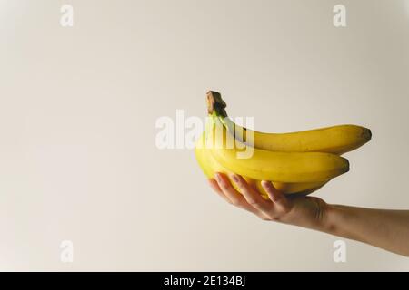 Gros plan sur la main d'une femme caucasienne inconnue qui détient des bananes devant le mur blanc - aliments biologiques sains frais espace de copie fruit concept Banque D'Images