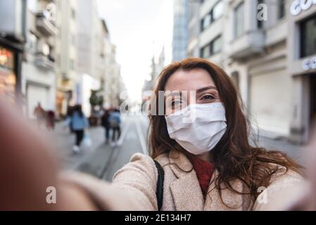 Belle fille portant un masque médical de protection et des vêtements tendance prend selfie avec un smartphone dans la rue. Nouveau concept de mode de vie normal. Banque D'Images