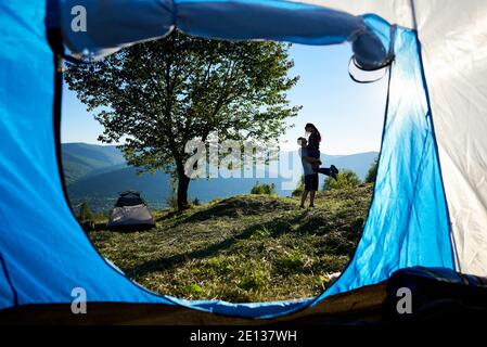 Vue arrière de deux jeunes randonneurs heureux homme et femme se reposant près du camping et grand arbre dans les montagnes le matin ensoleillé. Gars tenant une fille dans les mains. Vue de l'intérieur de la tente des touristes Banque D'Images