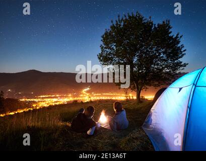 Vue arrière de jeunes touristes de couple se reposant au feu de camp à côté du camp et de la tente bleue, boire du café, profiter du ciel nocturne incroyable plein d'étoiles. En arrière-plan ciel étoilé, montagnes, arbre et ville lumineuse Banque D'Images