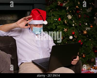 Un homme dans un chapeau de Noël et un masque médical souhaite une bonne année en ligne. Vacances d'hiver à la maison pendant la quarantaine. Banque D'Images