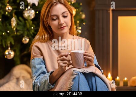 Femme dans des vêtements chauds et avec une tasse de thé près de la cheminée à la maison la veille de Noël. Concept de saison de chauffage Banque D'Images