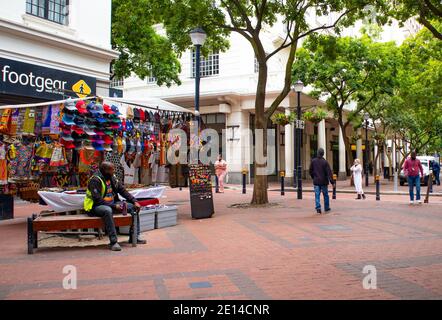 Cape Town, Afrique du Sud - 23/11/2020 marché occupé. Des vêtements colorés ainsi que des masques de visage suspendus dans un marché, la sécurité assis sur un banc Banque D'Images