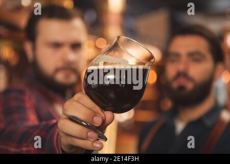 Gros plan de deux brasseurs examinant une délicieuse bière foncée dans un verre, travaillant à l'usine de production de bière. Accent sélectif sur une tasse de bière dans la ha Banque D'Images