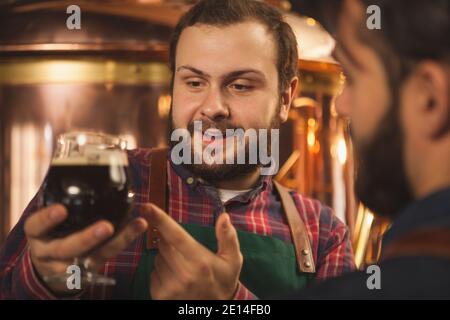 Gros plan d'un brasseur à bière à barbe souriant, parlant à son collègue pour examiner une bière sombre dans un verre. Deux brasseurs produisant de la bière artisanale à thei Banque D'Images