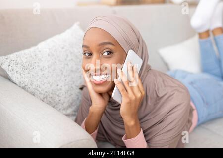 Jeune femme musulmane dans le hijab bavardant sur le téléphone portable, ayant une conversation agréable avec un ami tout en étant allongé sur le canapé à la maison Banque D'Images