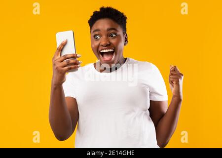 Femme africaine tenant un smartphone secouant les poings dans la joie, fond jaune Banque D'Images