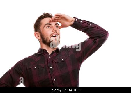 Beau barbu homme souriant souriant regardant loin avec sa main à ses yeux. Un homme séduisant et excité isolé sur un espace de copie blanc. Joyeux et émotif Banque D'Images