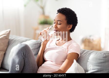 Hydratation pour une bonne santé concept. Jolie jeune femme noire ayant un verre d'eau claire sur le canapé à la maison Banque D'Images