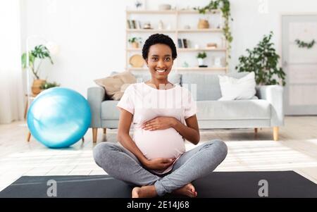 Concept de maintien de la forme pendant la grossesse. Jolie femme noire qui touche son ventre, assise sur un tapis de sport à la maison Banque D'Images