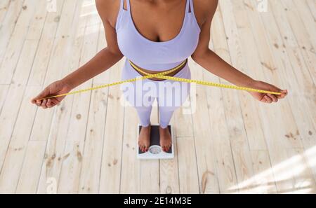 Gros plan d'une femme noire mince mesurant sa taille tout en étant debout sur une balance à la maison, au-dessus de la vue Banque D'Images