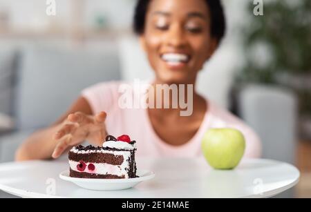 Concept de ventilation du régime. Jeune femme noire atteignant pour le gâteau au lieu de la pomme, en choisissant la nourriture de pourriels plutôt que saine à la maison Banque D'Images