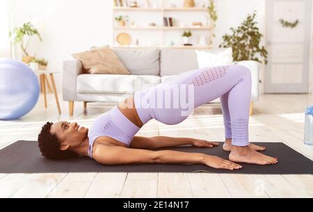 Femme noire sportive faisant des exercices sur le tapis de yoga, debout dans la moitié de pont pose à l'intérieur, vue latérale Banque D'Images