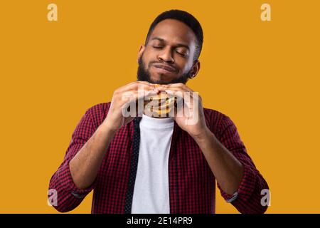 Happy African Man Eating Burger posant sur un fond de studio jaune Banque D'Images