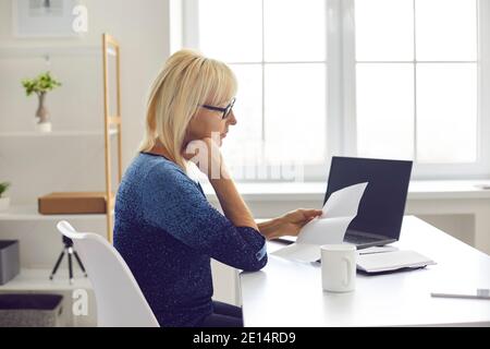 Femme sénior sérieuse assise à son bureau à la maison et lecture de la lettre de notification Banque D'Images