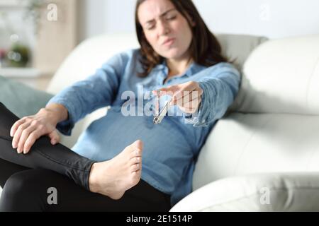 Femme enceinte stressée ayant des difficultés à couper ses ongles de pied sur un canapé à la maison Banque D'Images