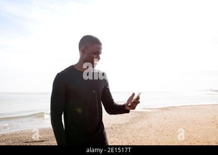 Le sportif noir sourit alors qu'il parle à son téléphone portable sur la plage avec un rétro-éclairage. Concept de sport et de santé. Banque D'Images