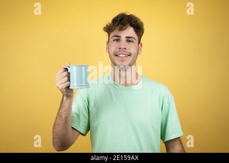 Beau homme portant un t-shirt vert décontracté sur fond jaune apprécier et boire une tasse de café Banque D'Images