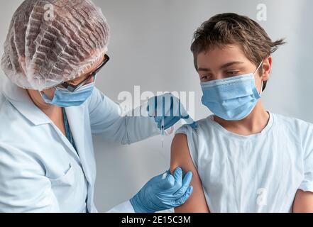 Un enfant anxieux qui fait le visage, peur de la seringue. Medic, médecin, infirmière, praticien de santé en robe blanche et masque facial vaccine l'adolescent. Nouveau Banque D'Images