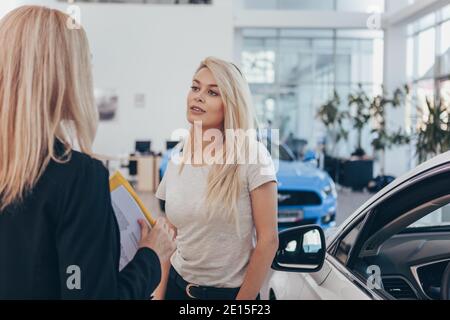 Belle femme achetant une nouvelle voiture à la concession, parlant à la vendeuse, espace de copie. Concessionnaire automobile professionnel aidant son client à choisir une nouvelle voiture Banque D'Images