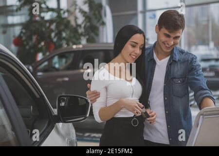 Jeune heureux couple magasiner à la concession automobile pour la nouvelle automobile. Belle femme et son beau mari gai choisissant l'auto à acheter. Homme achetant c Banque D'Images