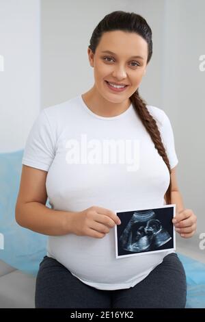 Portrait d'une femme enceinte charmante avec des cheveux foncés tenant la photo après le dépistage par ultrasons. Bonne femme en t-shirt blanc, assise sur un canapé médical et regardant la caméra. Banque D'Images