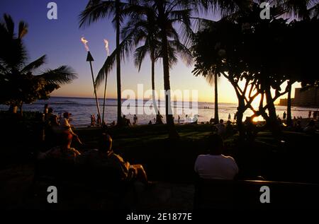 People seated on benches at Waikiki Beach at sunset with tiki torches lit Stock Photo