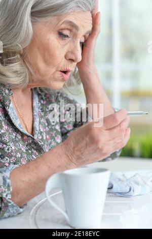 Femme sénior malade assise à la table à la maison avec un thermomètre Banque D'Images