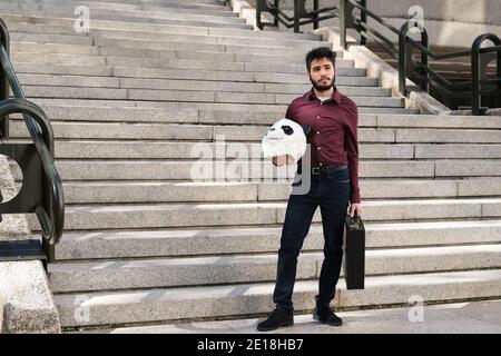 Jeune homme d'affaires avec un masque de tête de panda dans ses mains et une valise, debout sur les escaliers. Homme d'affaires bizarre. Banque D'Images