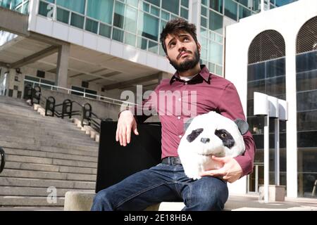 Jeune homme d'affaires avec un masque de tête de panda dans ses mains et une valise, assis sur un banc. Homme d'affaires bizarre. Banque D'Images