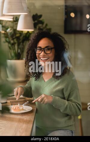 une femme afro-américaine joyeuse dans des lunettes tenant des couverts près de la tarte sur la plaque Banque D'Images