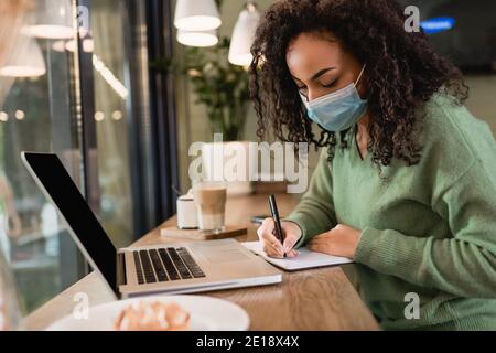 femme afro-américaine dans le masque médical écrivant dans le bloc-notes près ordinateur portable avec écran vierge sur la table Banque D'Images