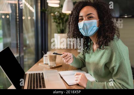 femme afro-américaine dans un masque médical tenant un stylo près d'un ordinateur portable et ordinateur portable avec écran vierge sur la table Banque D'Images