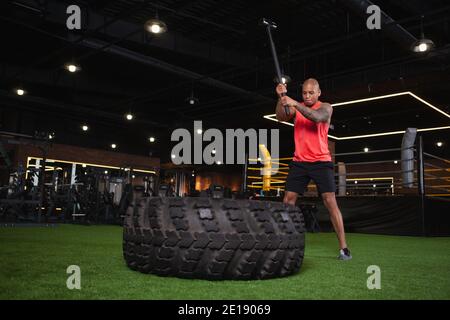 Prise de vue en longueur d'un puissant athlète africain de sexe masculin faisant de l'exercice de massette à la salle de gym. Un sportif musclé qui frappe un énorme pneu de roue avec de la hea Banque D'Images