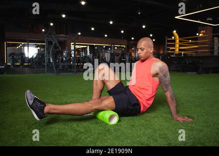 Attrayant jeune athlète africain masculin relaxant les muscles de jambe après l'entraînement de gym, en utilisant le rouleau en mousse. Homme athlétique et élégant se reposant après avoir fait de l'exercice, en utilisant Banque D'Images