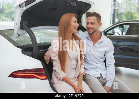 Heureux beau couple parlant, assis dans le coffre de leur nouvelle voiture à la concession. Couple achetant une nouvelle automobile Banque D'Images