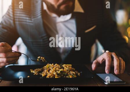 Homme barbu adapté qui vérifie le téléphone tout en mangeant de la nourriture dans un restaurant élégant. Banque D'Images