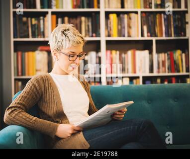 Magnifique fille blonde heureuse avec des lunettes lisant un livre tout en étant assis sur le canapé devant la bibliothèque. Banque D'Images