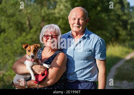 Couple de personnes âgées posant avec leur petit chien Jack Russell, la langue dehors. Femme la tient sur les mains, arbres flous et arrière-plan de la route de campagne Banque D'Images