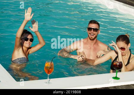 Groupe d'amis dansant dans l'eau à la fête de la piscine. Des jeunes joyeux qui se font boire un verre, rient joyeusement, profitent d'une chaude journée d'été au Swimmi Banque D'Images