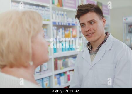 Jeune homme chimiste élégant parlant à son client masculin senior, travaillant à la pharmacie. Sympathique gai employé de pharmacie aider client âgé. M Banque D'Images