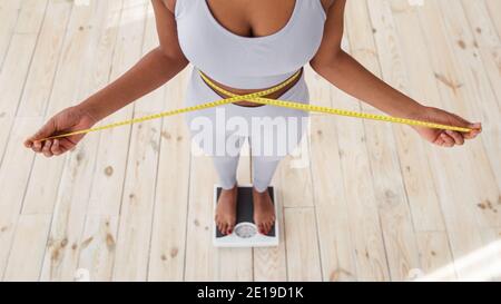 Au-dessus de la vue de la femme afro-américaine mesurant sa taille avec du ruban adhésif, debout sur des balances à l'intérieur, gros plan Banque D'Images