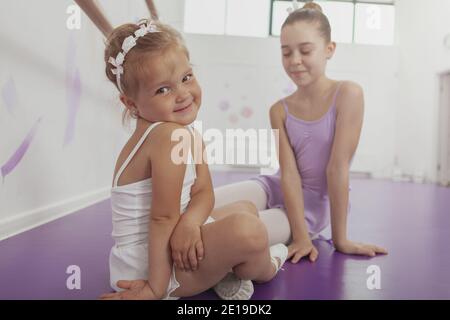 Adorable petite ballerine souriant à la caméra au-dessus de son épaule, se reposant après avoir pratiqué le ballet avec sa sœur aînée. Les filles charmantes se détendent Banque D'Images