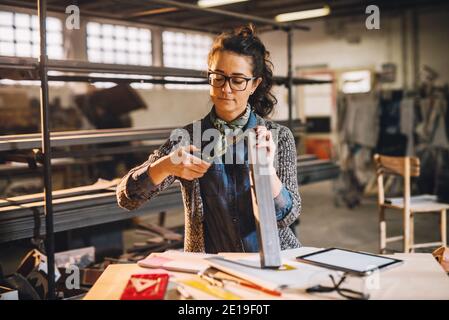 Vue rapprochée de la femme d'affaires motivée et professionnelle qui travaille dur et qui mesure un tuyau métallique avec un mètre ruban dans l'atelier de tissu ensoleillé. Banque D'Images