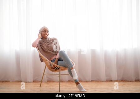 Jolie femme noire dans hijab assis sur un fauteuil confortable et en regardant de côté l'espace vide à l'intérieur Banque D'Images