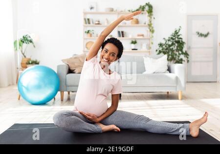 Grossesse active. Future maman noire souriante s'étirant les jambes, faisant son entraînement de yoga du matin sur le tapis à la salle de gym à la maison Banque D'Images