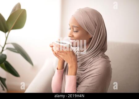 Belle dame noire dans le hijab traditionnel odeur de café, appréciant l'arôme magique avec les yeux fermés, à l'intérieur Banque D'Images