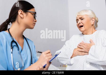 femme âgée touchant la poitrine tout en souffrant d'asthme attaque près d'une infirmière asiatique écrivant sur le presse-papiers, flou de premier plan Banque D'Images