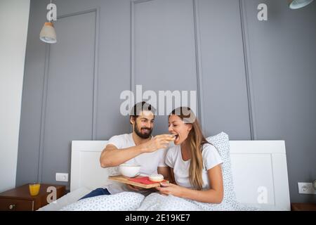 Homme joyeux barbu donnant une belle jeune fille un peu d'un petit déjeuner au lit. Banque D'Images